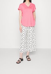 Rosa kurzärmlige Bluse mit Kragen, kombiniert mit weißen, weit geschnittenen Hosen mit schwarzen Punkten. Schwarze Sandalen mit Blockabsatz und Riemchen.