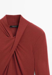 Červený top s drapedovým, zkrouceným výstřihem a dlouhými rukávy. Měkká textura, s viditelnou značkovou etiketou "Massimo Dutti" na límci.