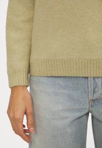 Ljus olivgrön stickad tröja med ribbat upptäck, bärs över blekta blå denimjeans. Närbild av hand som vilar på den denimen.