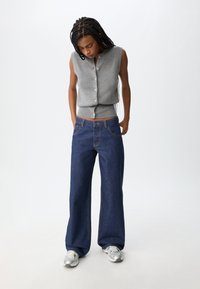 Šedý top bez rukávů s knoflíky, který je spárován s modrými širokými džíny z denimu s klasickým pětiokým designem a světlým prošíváním.