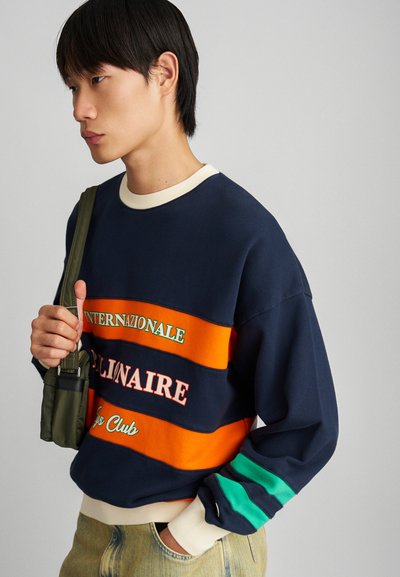 Sweatshirt bleu marine avec des rayures orange et vert, comportant des motifs textuels. Col et manches côtelés ; coupe décontractée. Porté avec un petit sac.