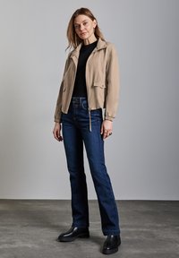Béžová zipová bunda s elastickými manžetami, nošená přes černý top. V kombinaci s tmavě modrými rozšířenými džíny a černými kotníkovými botami.