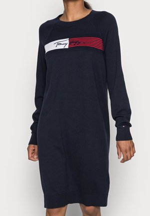 Granatowa dzianinowa sukienka z długim rękawem i okrągłym dekoltem, z panelem na piersi w czerwono-białe paski oraz logo w stylu kursywy.
