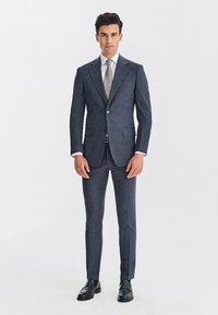 Ciemnoniebieski garnitur na miarę z dwoma guzikami, klapami z wycięciem oraz dopasowanymi spodniami. Biała koszula i wzorzysty krawat, uzupełnione czarnymi eleganckimi butami.