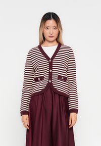 Γυναίκα που φοράει μια μπορντό και λευκή ριγέ cardigan με κουμπιά πάνω από ένα λευκό μπλουζάκι και μπορντό φούστα, στέκεται μπροστά σε έναν απλό λευκό φόντο.