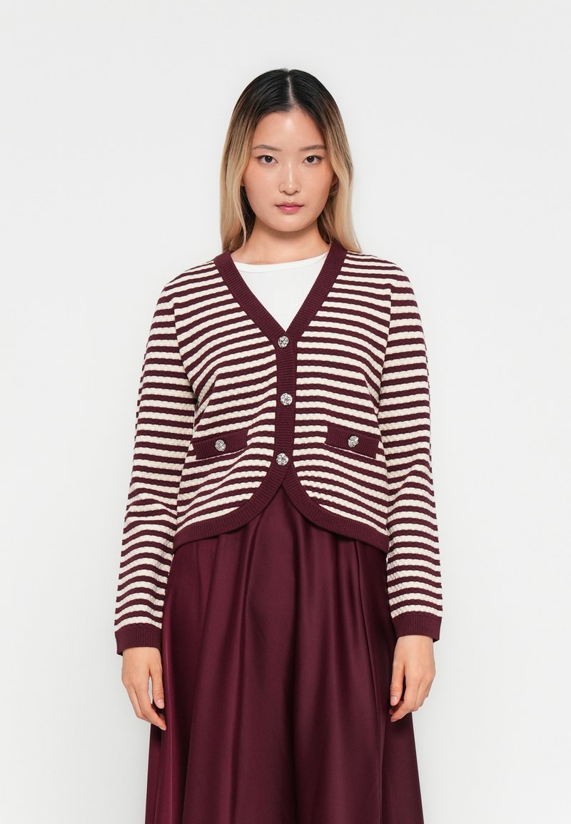 Γυναίκα που φοράει μια μπορντό και λευκή ριγέ cardigan με κουμπιά πάνω από ένα λευκό μπλουζάκι και μπορντό φούστα, στέκεται μπροστά σε έναν απλό λευκό φόντο.