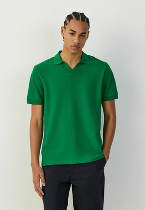 Junger Mann mit geflochtenem Haar, der ein hellgrünes kurzärmeliges Poloshirt und schwarze Hosen trägt, vor einem schlichten weißen Hintergrund steht.