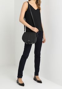 Bolso bandolera de cuero negro con forma curva, correa delgada y herrajes plateados. La modelo lleva jeans oscuros y una blusa negra, posando frente a una pared clara.