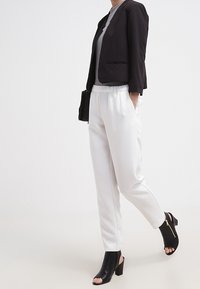 Femme portant un blazer noir, un haut gris, un pantalon blanc à taille élastique et des bottines à talons ouvertes noires, marchant devant un fond uni.