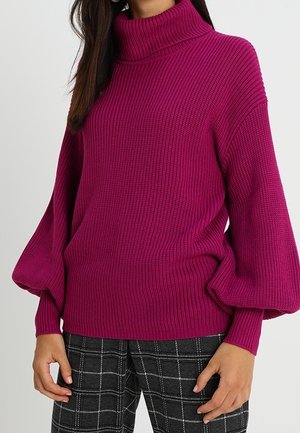 Femme portant un pull à col roulé côtelé magenta avec des manches bouffantes et un pantalon à carreaux noir, debout devant un fond clair uni.