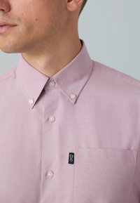 Cămașă roz deschis cu nasturi, cu guler, prevăzută cu un buzunar pe partea stângă a pieptului și logo brodat negru. Textură netedă a materialului și design clasic.