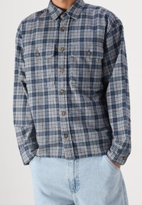 Camisa de franela a cuadros azul y gris con cierres de botones, dos bolsos en el pecho y cuello tipo solapa. La tela tiene una textura suave.