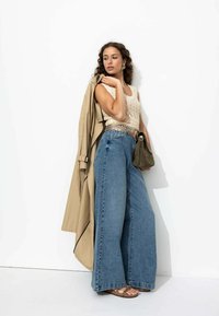 Trench-coat beige, haut en crochet crème, jeans en denim bleu à jambes larges, ceinture métallique, et sandales brunes à motifs. Sac à main marron en accent.
