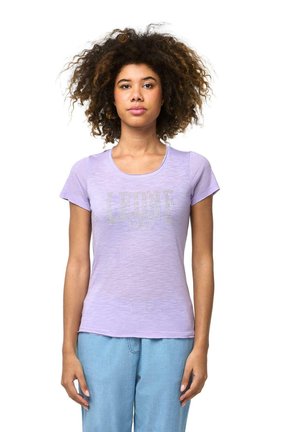 Une femme aux cheveux bouclés se tient devant un fond blanc. Elle porte un T-shirt couleur lavande avec un logo argenté et un jean bleu clair.