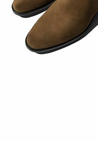 Botas de tobillo de ante marrón con punta afilada y suela de goma negra. Textura suave con líneas limpias y detalle de costura sutil.