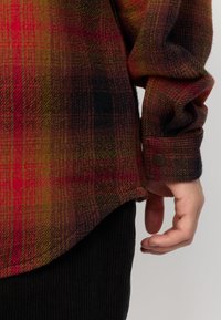 Rood, geel en zwart geruite flanellen shirt met een afgeronde zoom, knoopmanchetten en een zachte textuur. Draag het over zwarte corduroybroek.