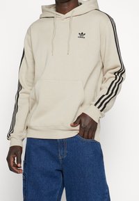 Beige hoodie med svart Adidas-logga, dragskohuva och tre svarta ränder längs ärmarna. Har en framfickan i kängurudesign.