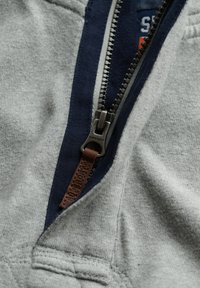 Grijs sweatshirt stof met een marineblauw accent en een metalen rits met een bruine leren trekker met het label "Superdry."