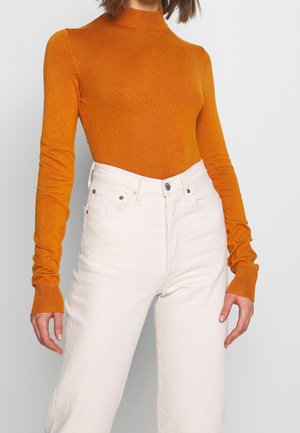 Pull à col roulé orange en tricot doux, associé à un jean blanc taille haute avec fermeture à boutons et détails de poches subtils.