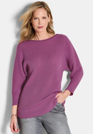 Donna con capelli biondi che indossa un maglione viola a costine e jeans grigi con strass, mentre posa con una mano in tasca su uno sfondo bianco.