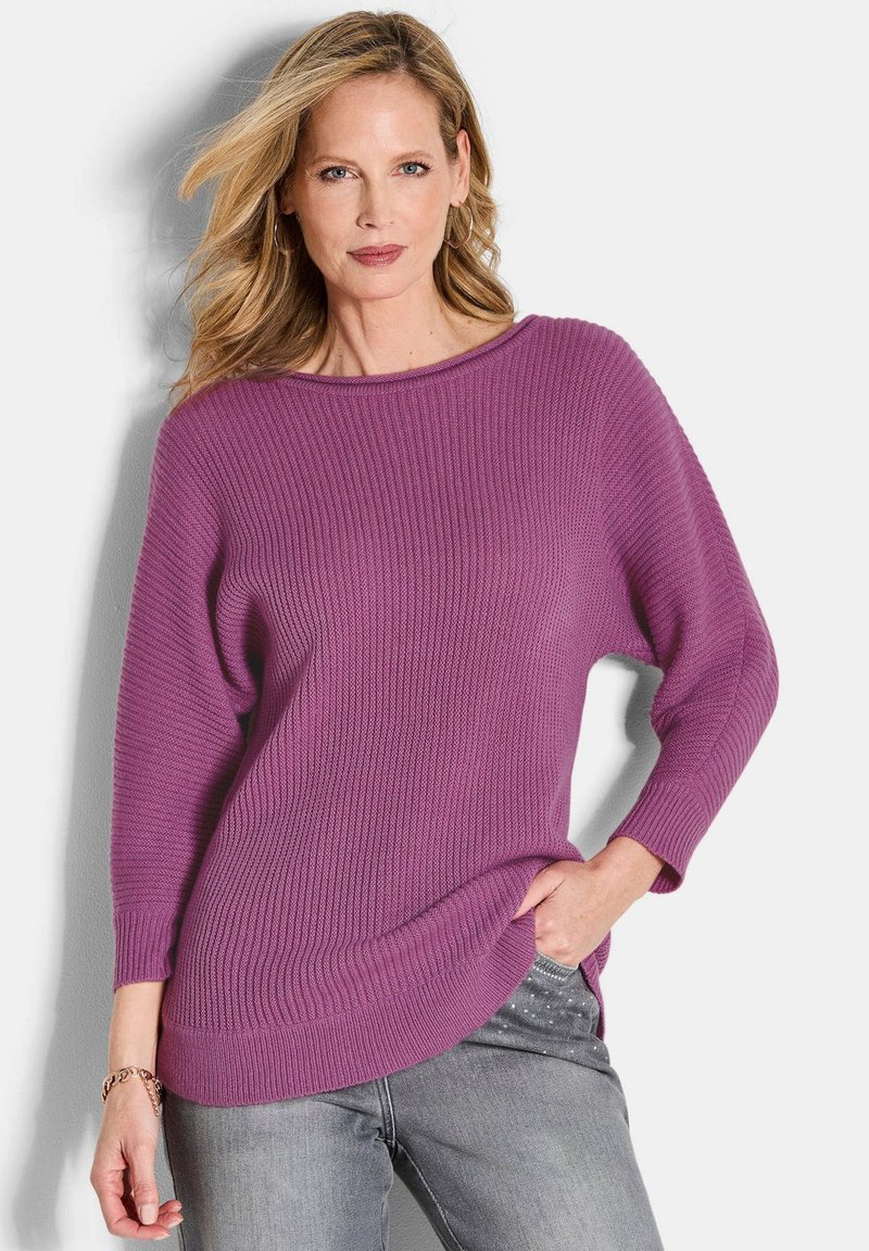 Frau mit blonden Haaren, die einen lila gerippten Pullover und graue Jeans mit Strass trägt, posiert mit einer Hand in der Tasche vor einem weißen Hintergrund.