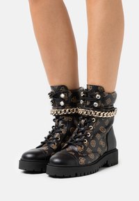 Bottes de combat noires avec un motif doré, design à lacets, quincaillerie argentée et semelle noire épaisse. Dotées d'une chaîne décorative autour de la cheville.