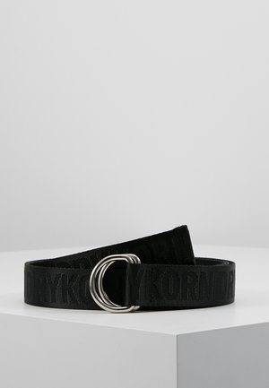 Ceinture en tissu noir avec texte embossé et boucle double D en argent, présentée sur une surface blanche devant un fond gris.