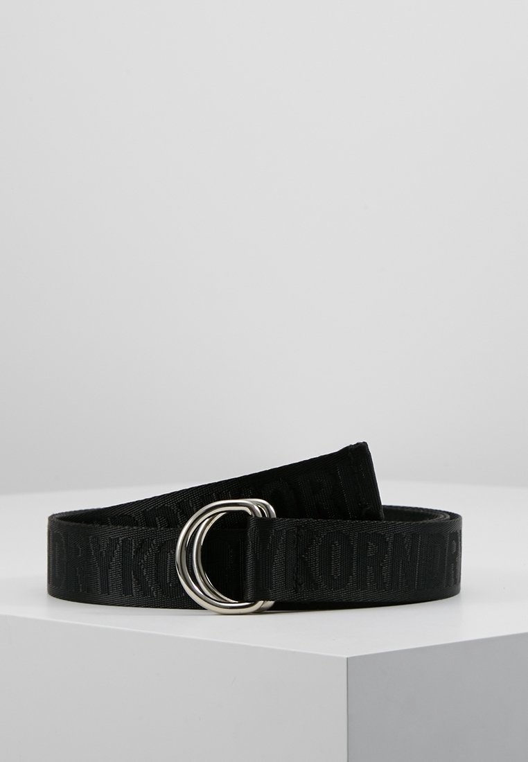 Ceinture en tissu noir avec texte embossé et boucle double D en argent, présentée sur une surface blanche devant un fond gris.