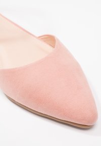 Des ballerines en suede rose avec un bout pointu et des côtés ouverts, présentant une texture lisse et une doublure intérieure beige, sur un fond blanc.