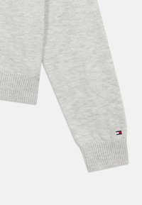 Gaiši pelēks adīts džemperis ar ribotiem piedurknēm un nelielu Tommy Hilfiger karoga logo tuvu plaukstas locītavai uz baltas fona.