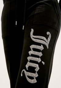 Melnas veliūra sporta bikses ar sudraba spīdīgu "Juicy" logo uz augšstilba, ar aukliņu jostasvietā un mīkstu tekstūru.
