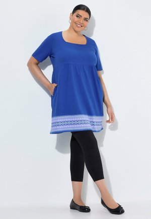 Mujer sonriente con túnica azul de borde estampado, leggings negros y zapatos planos negros, de pie contra un fondo blanco con una mano en el bolsillo.