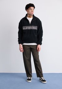 Sort fleece pullover med en kvart lynlås, der har et gråt "Quiksilver" logo på brystet, parret med olivengrønne bukser og sorte sneakers.