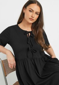 Mustat puuvillamekko lyhyillä hihoilla, jossa on pyöreä pääntie ja solmukkeita edessä. Laiska hame ja pehmeä rakenne.