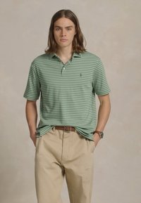 Camisa polo verde claro com riscas horizontais brancas, pala de dois botões e logótipo bordado, combinada com calças beges. Ajuste simples e descontraído.