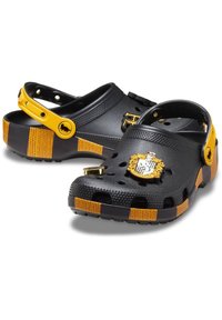 Schwarze Crocs mit gelben Akzenten; verfügen über eine strukturiertes Obermaterial, Belüftungsöffnungen und ein goldenes Emblem an der Vorderseite. Verstellbarer Riemen auf der Rückseite.