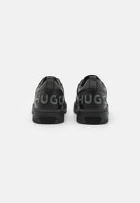 Sneakers in pelle nera con il logo "HUGO" in materiale lucido sulla parte posteriore, dotati di suole in gomma nera texturizzata.