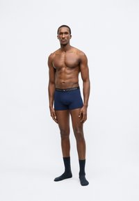 Meresinised bokserpüksid tekstuurse mustriga ja musta värvusega vöökohaga, millel on kuldsete tähtedega kiri "TOMMY HILFIGER", kantud tumedate sokkidega.