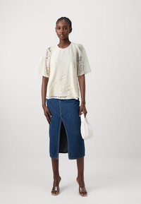 Top in pizzo floreale crema con maniche corte, abbinato a una gonna di jeans con uno spacco frontale. Il modello tiene una pochette bianca e indossa sandali trasparenti.