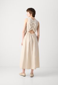 Beige sleeveless dress with a scoop back and cutout detail. Features a cinched waist, airy fabric, and falls to mid-calf. Worn with beige shoes.