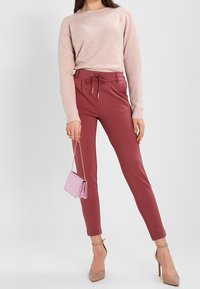 Kobieta w jasnoróżowym swetrze, spódnicy w kolorze mauve z wysokim stanem i wiązanym paskiem, w cielistych szpilkach, trzymająca małą różową torebkę na łańcuszku.