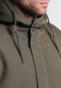 Veste à capuche vert olive avec fermeture éclair et pression, cordons ajustables et tissu lisse et texturé.