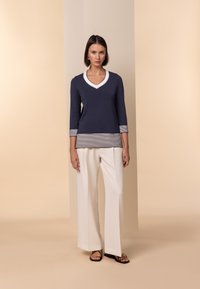 Maglione a V blu navy con orlo e polsini a righe, abbinato a pantaloni bianchi a gamba larga e sandali neri. Tessuto morbido e vestibilità rilassata.