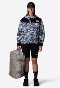 North Face abstract-patterned jacket in blue and black, black shorts, carrying a gray soft duffel bag, wearing black shoes and cap.