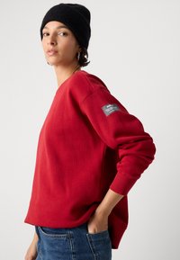 Frau trägt einen roten Langarm-Sweater mit einer schwarzen Mütze und blauen Jeans, sie steht mit einer Hand in ihrer Tasche und schaut zur Seite.