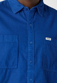 Blå kord skjorta med knapplist, två bröstfickor och en liten logotags. Har textilerad väv och en klassisk kragdesign.