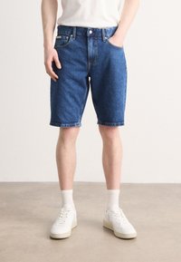 Mørkeblå denimshorts med klassisk design, som har sidelommer og rett snitt, kombinert med hvite sneakers og ankelsokker.