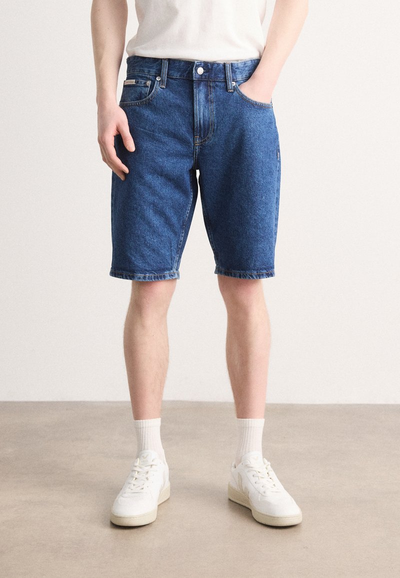 Mørkeblå denimshorts med klassisk design, som har sidelommer og rett snitt, kombinert med hvite sneakers og ankelsokker.