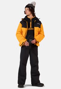 Donsjack in oranje en zwart, fleece binnenlaag, zwarte broek, een gestreepte beanie met een pompon op, houdt zwarte wanten vast.
