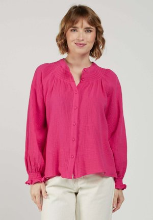 Femme portant un chemisier rose à manches longues avec des boutons, en tissu texturé et des poignets froncés, associé à un pantalon clair.